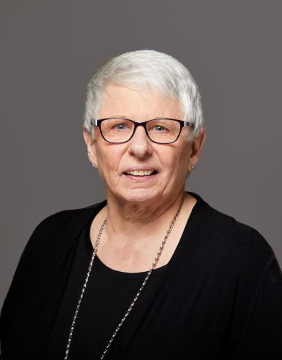 Headshot of  woman with black-framed glasses and short grey hair, wearing a black top and long silver necklace