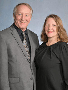 Professional photo of Daniel and Debbie, older man smiling wearing a gray suit and older women smiling with brown hair wearing a black blouse