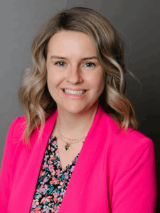 Professional photo of a female general manager with shoulder-length blonde-brown hair wearing a colorful floral top and a bright pink blazer