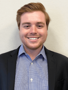 Professional photo of a young male sales and account manager with short red hair wearing a blue plaid button-down shirt and a black blazer