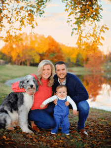 Professional photo of the Tibbetts family, in a field of autumn leaves and a pond in the back. Rachel is smiling, wearing an orange blouse, hugging their big dog, Matt is kneeled down, smiling, wearing a blue sweater and jeans, holding their baby boy wearing blue overalls.